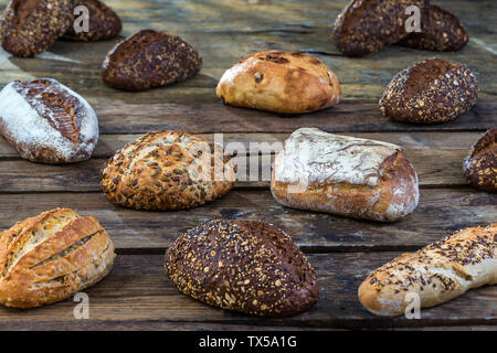Several small multi grain different shaped bread sprinkled with whole sunflower seeds, flax and sesame seeds and wheat and barley spikes on old brown wooden table Stock Photo