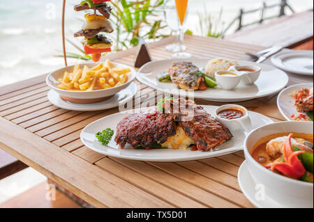 Variety of food, roasted pork ribs, beef steak, seafood and spicy soup on dining table in tropical sea Stock Photo