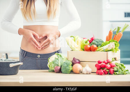 Woman with sports figure on her belly shows heart shape in home kitchen with wooden box full of organic vegetable. Stock Photo