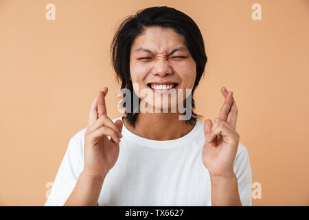 Photo closeup of asian guy 20s wearing white t-shirt keeping fingers crossed and making a wish isolated over beige background Stock Photo