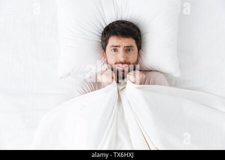 Image of a scared handsome young man in the morning under blanket hide in bed. Stock Photo