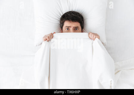 Image of a scared handsome young man in the morning under blanket hide in bed. Stock Photo