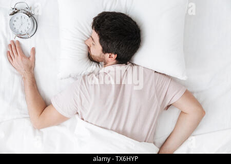 Image of a sleeping young man in the morning under blanket in bed lies with alarm clock. Stock Photo