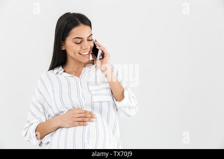 Smiling young pregnant businesswoman standing isolated over white background, talking on mobile phone Stock Photo