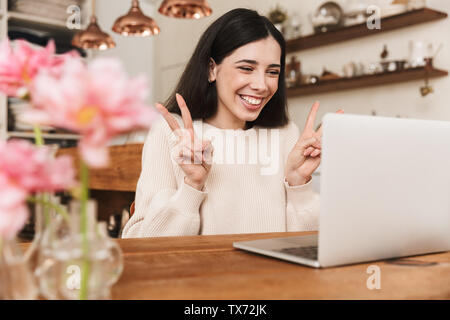 Smiling young woman sitting at the kitchen and using laptop computer, having video call Stock Photo