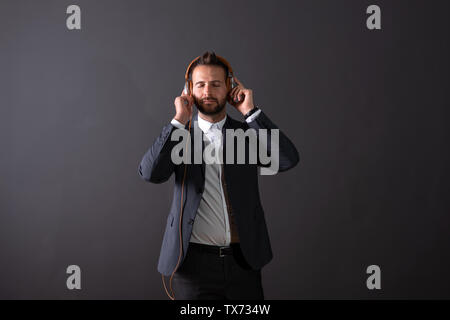 A handsome young businessman in a suit listening to music and enjoying it in front of a dark grey background in a studio. Stock Photo
