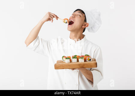 Image of happy chinese chief man in white cook uniform holding plate and eating sushi set with chopsticks isolated over white background Stock Photo