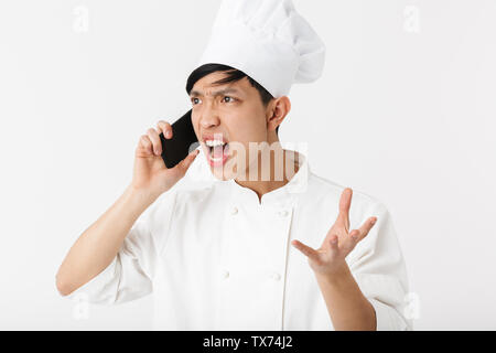 Image of angry chinese chief man in white cook uniform and chef's hat talking on mobile phone isolated over white background Stock Photo