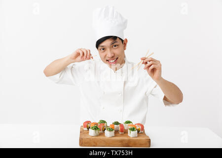 Image of pleased chinese chief man in white cook uniform eating sushi set with chopsticks isolated over white background Stock Photo