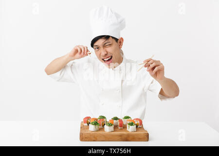 Image of playful asian chief man in white cook uniform eating sushi set with chopsticks isolated over white background Stock Photo