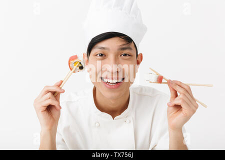 Image of funny asian chief man in white cook uniform eating sushi set with chopsticks isolated over white background Stock Photo