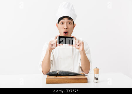 Image of surprised japanese chief man in white cook uniform holding smartphone while fillet raw fresh fish isolated over white background Stock Photo