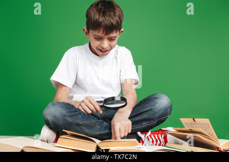 Cute little boy with freckles studying, sitting with stack of books over green background, holding magnifying glass Stock Photo