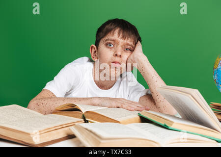 Cute confused little boy with freckles studying, sitting with stack of books over green background Stock Photo