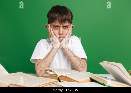 Cute confused little boy with freckles studying, sitting with stack of books over green background Stock Photo