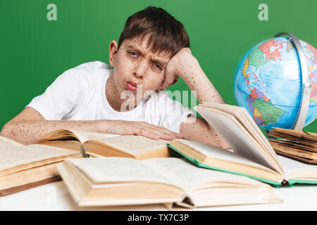 Cute confused little boy with freckles studying, sitting with stack of books over green background Stock Photo