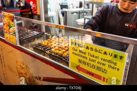 Dotonbori, Osaka, Japan - November 21, 2018: Woman cooking takoyaki behind counter with direction to buy ticket. Stock Photo