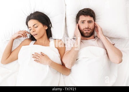 Image of a displeased man covering ears near his woman sleeping in bed under blanket. Stock Photo