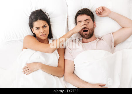 Image of a displeased woman covering nose of her man sleeping in bed under blanket. Stock Photo