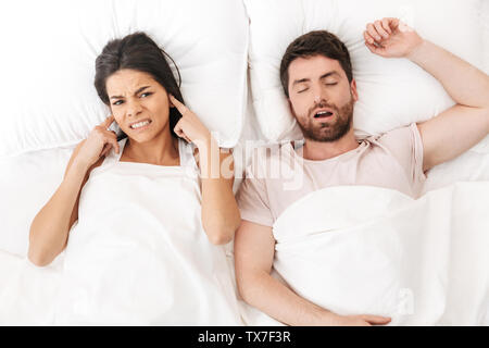 Image of a displeased woman covering ears near her man sleeping in bed under blanket. Stock Photo