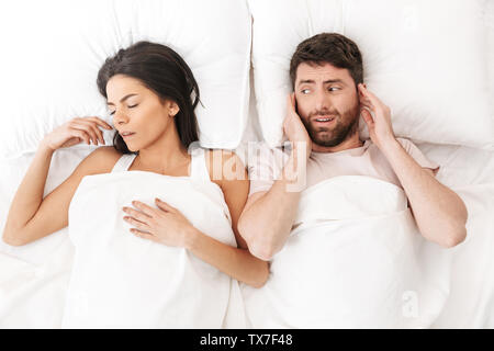 Image of a displeased man covering ears near his woman sleeping in bed under blanket. Stock Photo