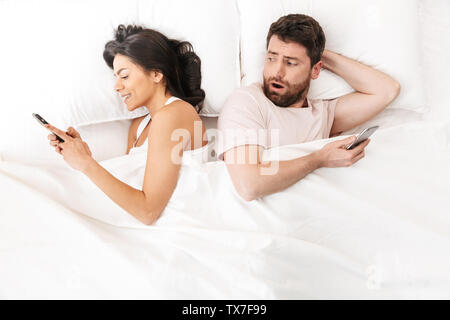 Image of a young loving couple lies in bed under blanket using mobile phones. Confused man looking at woman's phone. Stock Photo