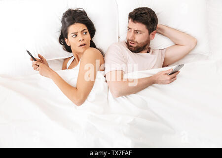 Image of a young confused loving couple lies in bed under blanket using mobile phones looking to each other. Stock Photo