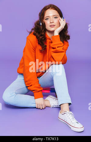 Portrait of a smiling young redheaded woman listening to music with headphones while sitting isolated over violet background Stock Photo