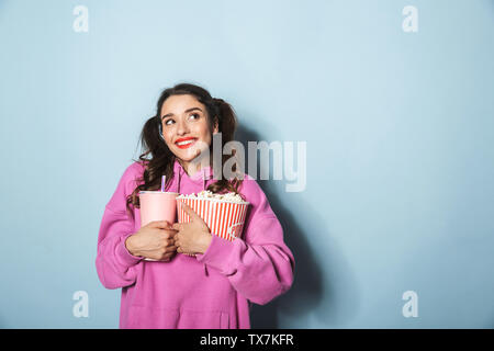 Portrait of happy young woman with two ponytails holding soda beverage in plastic cup and popcorn bucket isolated over blue background in studio Stock Photo