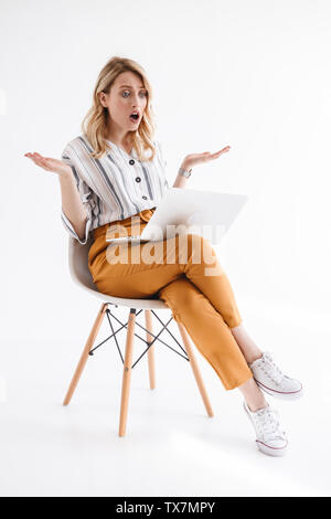 Portrait of shocked girl wearing casual clothes looking at laptop and throwing up hands while sitting in chair isolated over white background in studi Stock Photo