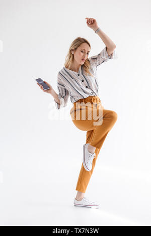 Full length photo of european woman dancing and holding mobile phone while listening to music via bluetooth earpod isolated over white background Stock Photo