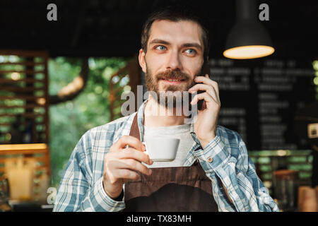 Portrait of attractive barista guy wearing apron drinking coffee and talking on cellphone in street cafe or coffeehouse outdoor Stock Photo
