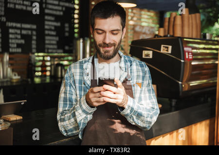 Portrait of caucasian barista guy wearing apron smiling and typing on mobile phone in street cafe or coffeehouse outdoor Stock Photo