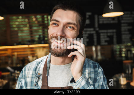 Portrait of joyful barista guy wearing apron smiling and talking on cellphone in street cafe or coffeehouse outdoor Stock Photo