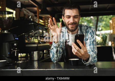 Photo of happy barista guy wearing apron smiling while using smartphone in cafe or coffeehouse outdoor Stock Photo