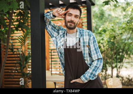 Portrait of young barista man wearing apron smiling at camera while working in street cafe or coffeehouse outdoor Stock Photo