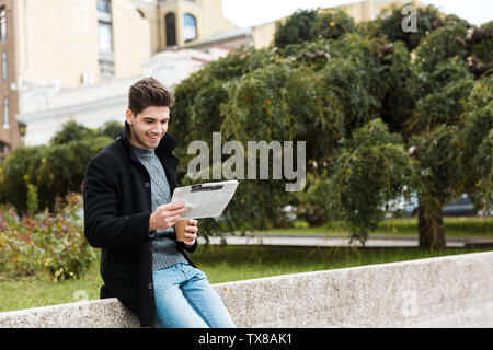 Photo of european man 30s wearing jacket reading newspaper and drinking takeaway coffee while walking through city park Stock Photo