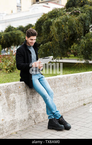 Photo of smiling man 30s wearing jacket reading newspaper and drinking takeaway coffee while walking through city park Stock Photo