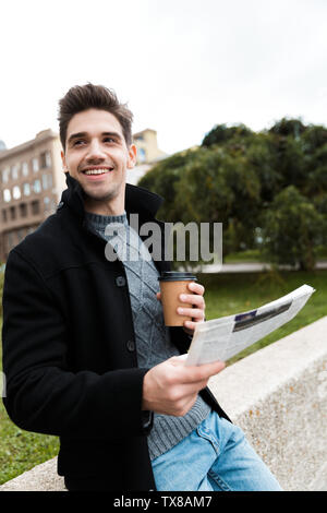 Photo of handsome man 30s wearing jacket reading newspaper and drinking takeaway coffee while walking through city park Stock Photo