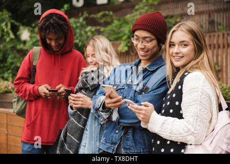 Group if cheerful multiethnic friends teenagers spending fun time together outdoors, standing, using mobile phones Stock Photo