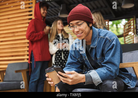 Smiling asian teenager sitting outdoors, holding takeaway coffee cup, using mobile phone Stock Photo