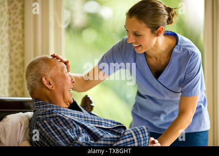 Female nurse leans over to place a hand on the forehead of a senior man lying in a bed. Stock Photo