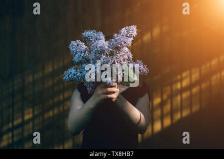 Girl holding lilac bouquet in front of face. Child enjoying flowers outdoors in front of wall. Kid hiding behind bunch of violet flowers. Summer and spring flora. Young person with gift in hands Stock Photo