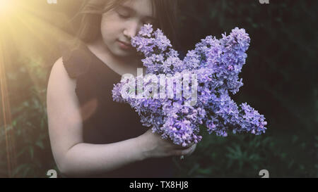 Cute little girl smelling lilac. Person holding flowers on dark natural background. Child enjoying bouquet outside. Going to park, forest in summer and spring. Connection with nature idea Stock Photo