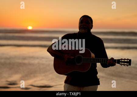 Happy senior man playing an acoustic guitar on a beach at sunset. Stock Photo