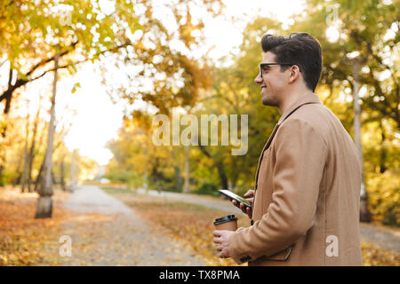 Portrait in profile of stylish young man wearing coat typing on smartphone and smiling while walking in autumn park Stock Photo
