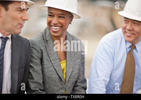 Three smiling engineers wearing suits and hardhats having a cheerful business discussion at a construction site. Stock Photo
