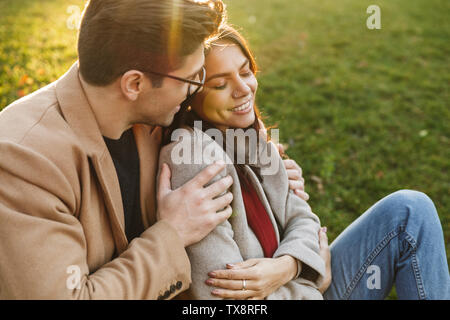Image of relaxed caucasian couple man and woman 20s smiling and hugging while sitting on grass in park Stock Photo