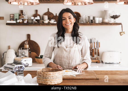 Portrait of smiling brunette woman 30s wearing apron cooking pastry with flour and eggs in kitchen at home Stock Photo