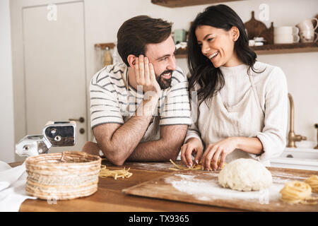 Portrait of positive couple man and woman 30s wearing aprons making homemade pasta of dough while cooking together in kitchen at home Stock Photo